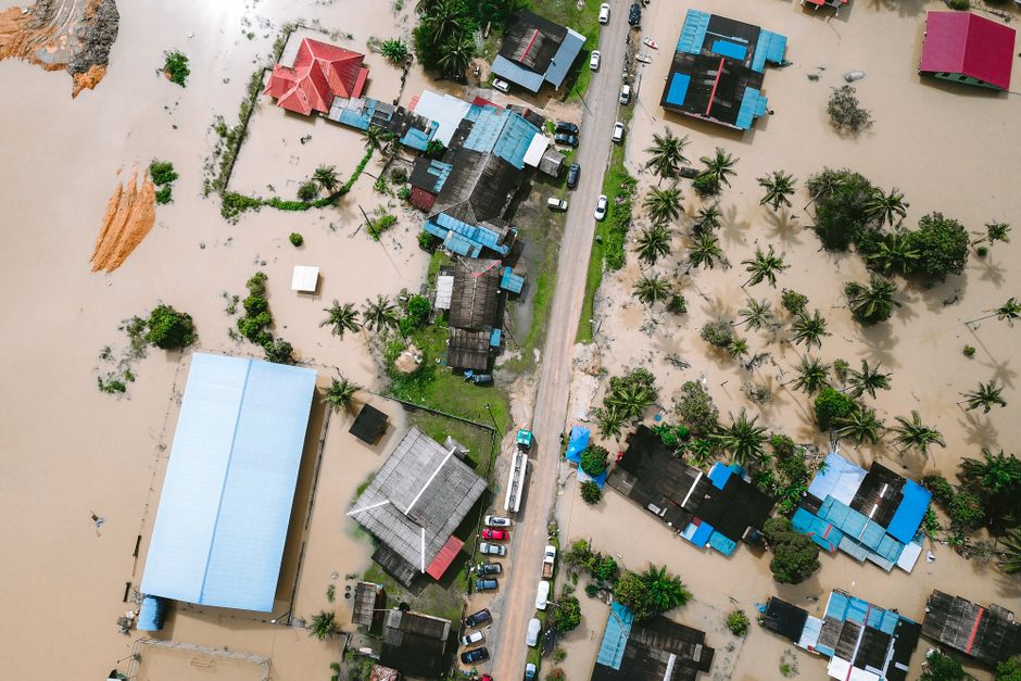 Aerial view of a residential area on Church Road in Upton, showing houses with tiled and metal roofs, some flooded with muddy water covering the surrounding grounds and roads. The flooded areas extend