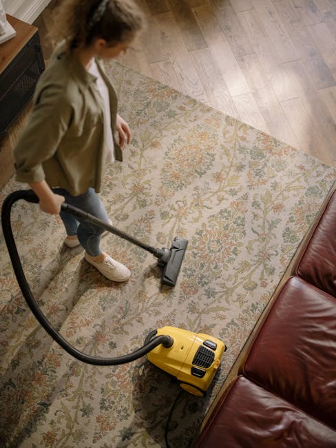 A person is vacuuming a decorative, patterned area rug with a yellow carpet cleaner in a residential living room. The individual is wearing beige shoes and light-colored pants, holding a black vacuum