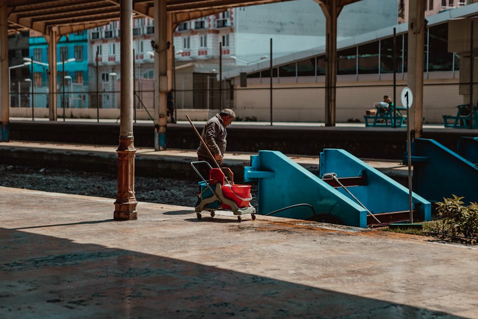A person wearing casual clothing and a face mask is performing surface cleaning in a dimly lit indoor space, using a long-handled floor tool to clean a carpeted area. The room features a large project